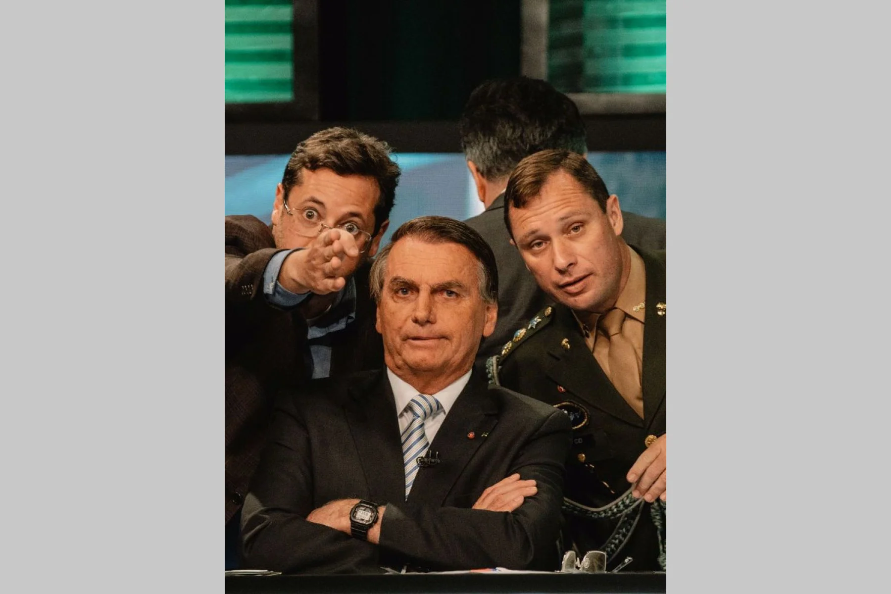 Jair Bolsonaro z założonymi rękami, za nim dwóch mężczyzn, jeden wskazuje, drugi w mundurze.
