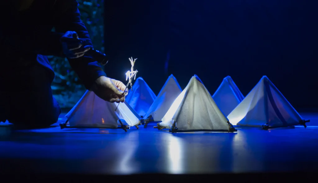 Osoba trzymająca małą kukiełkę nad rzędem miniaturowych białych namiotów na scenie teatralnej, oświetlonej niebieskim światłem.