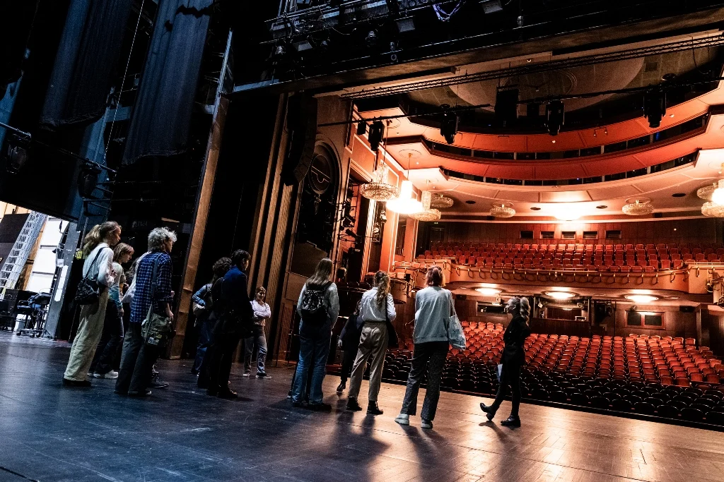 Grupa osób stojąca na dużej scenie w Sali Bogusławskiego, patrząca w kierunku rozświetlonej widowni i słuchająca edukatorki teatralnej.
