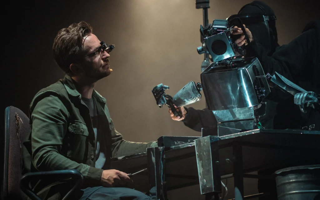 Mężczyzna w kurtce siedzi na fotelu przy metalowym stole. Patrzy na mechanicznego robota.