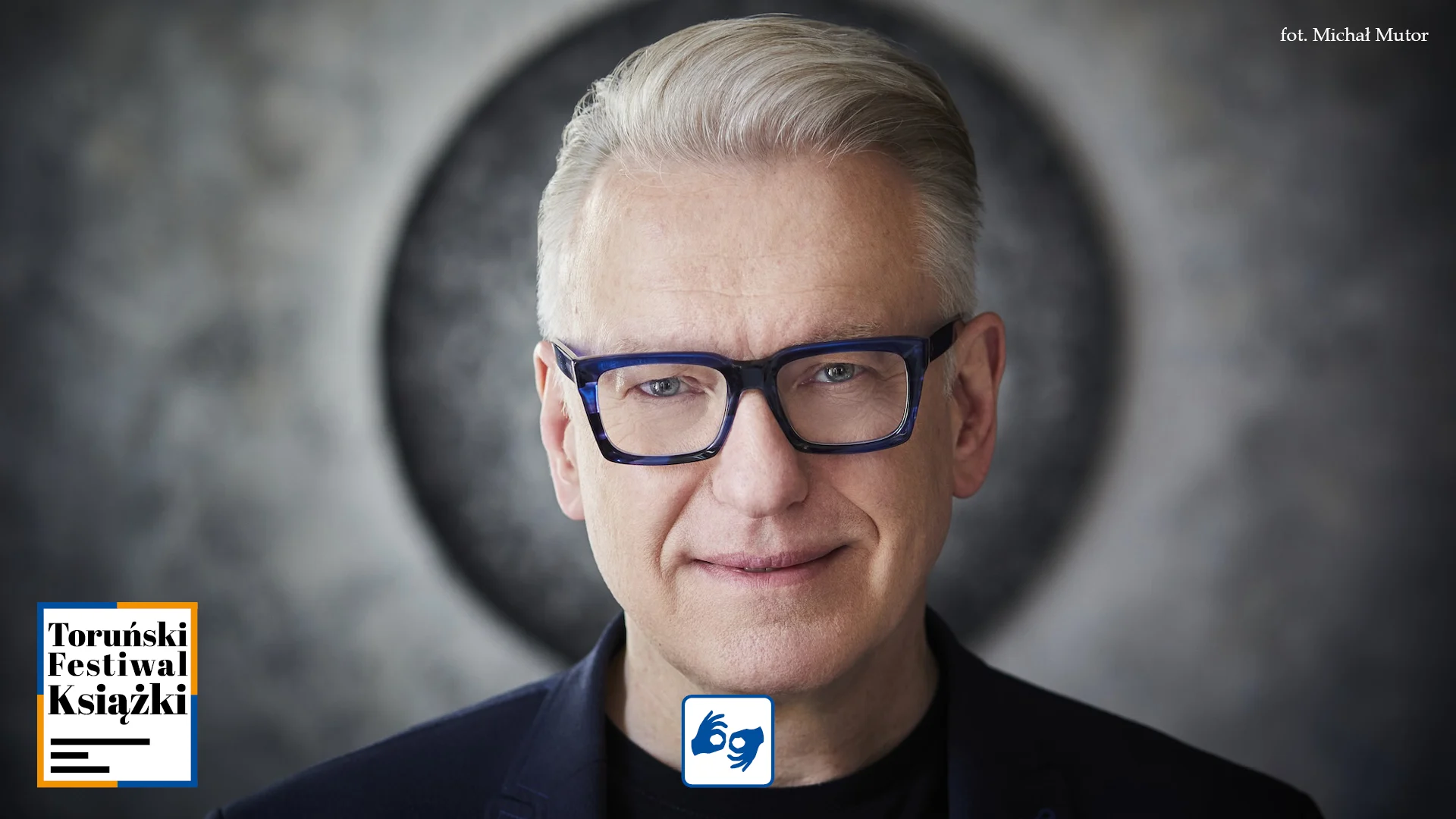 starszy mężczyzna z siwymi włosami elegancko ułożonymi na bok i w granatowych okularach, lekko się uśmiecha. Jest to Mariusz Szczygieł. Na dole po środku mały piktogram PJM